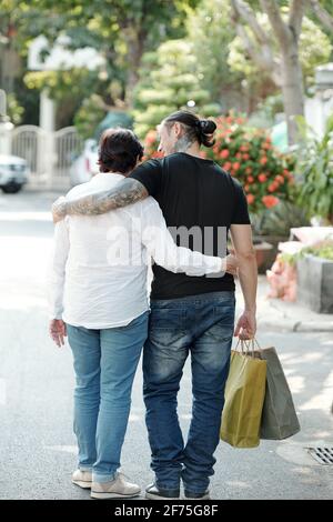 Uomo che cammina per strada con la madre più anziana e che porta le sue borse pesanti per lo shopping Foto Stock