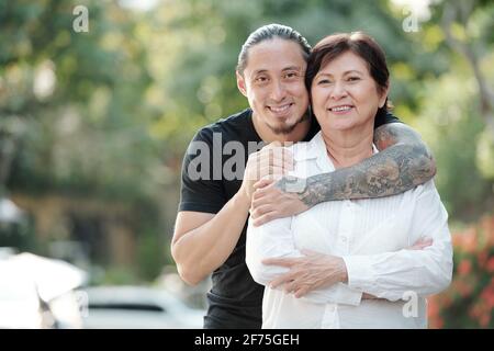 Ritratto di bell'uomo adulto di razza mista che abbracciava la madre matura da dietro e sorridendo alla fotocamera quando sono in piedi all'aperto Foto Stock