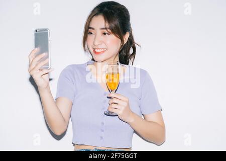 Giovane donna asiatica che beve vino e usa lo smartphone sul bianco sfondo Foto Stock