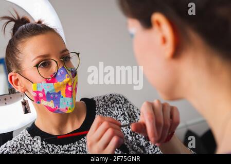 Primo piano di un estetista che indossa una colorata maschera protettiva durante l'applicazione make up su bella giovane donna in sedia Foto Stock