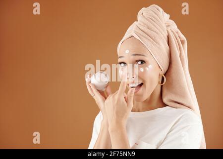 Felice giovane donna eccitata con asciugamano sulla sua testa che si applica lozione idratante dopo la presentazione mattutina Foto Stock