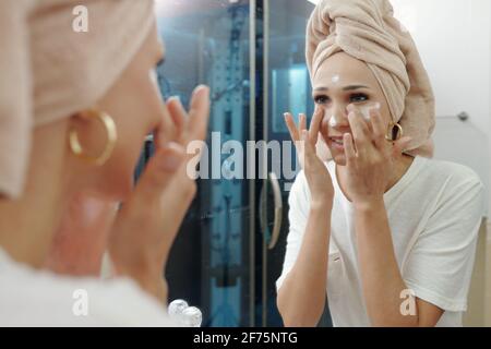 Donna giovane sorridente che guarda lo specchio quando si applica l'anti-invecchiamento idratante sul viso Foto Stock