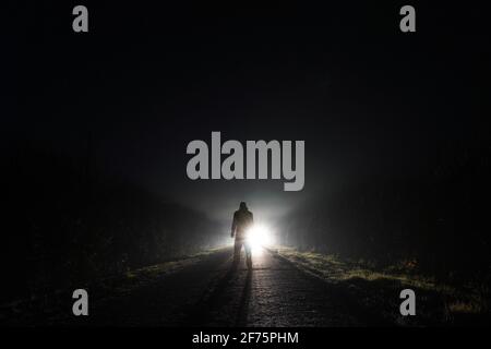 I fari dell'automobile illuminano indietro un uomo su una strada di campagna foggy spooky di notte. Foto Stock