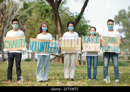 Gruppo di volontari in maschere protettive in piedi all'aperto con cartelli chiedere alle persone di unirsi a un'organizzazione benefica Foto Stock