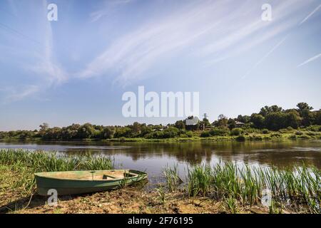 Vecchia barca solitaria sulla riva del fiume. Erba verde alta cresce. Sullo sfondo c'è un cielo blu e alberi bassi. Foto Stock
