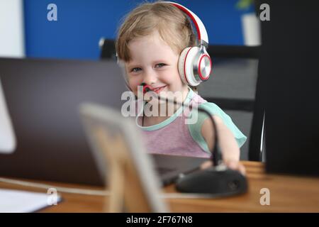 Piccola ragazza sorridente al tavolo da lavoro con un computer portatile in le cuffie sono accanto al microfono Foto Stock