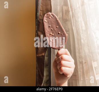 Una mano del bambino tiene un gelato di cioccolato su un bastone, un popsicle con noci sullo sfondo di un interno domestico in una giornata di sole. Foto Stock