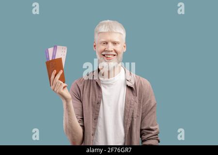 Concetto di voli economici. Felice uomo albino con passaporto e biglietti, in piedi isolati su sfondo turchese studio Foto Stock