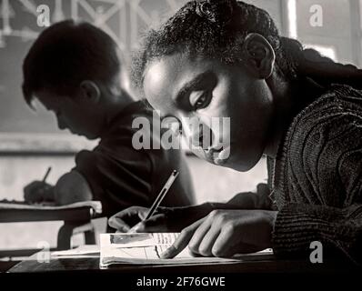 Junior ragazza classe scrivania scrittura scuola scolastica nero africano 9-11 anni di scrittura in classe alla sua scrivania con ragazzo biondo & la geometria della scuola proietta una lavagna dietro l'immagine del B&W. Foto Stock