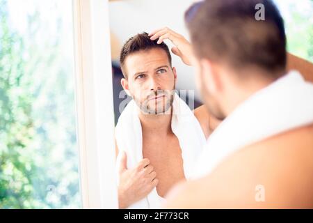 Primo piano di un bel giovanotto con asciugamano nel collo mentre fa i capelli nello specchio del bagno. Foto Stock