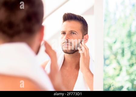 Primo piano di un bel giovane uomo con asciugamano nel collo ammirando il volto nello specchio del bagno. Foto Stock