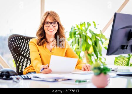 Foto di felice donna d'affari matura seduto alla scrivania dell'ufficio e facendo un po' di lavoro mentre lavora sul computer in ufficio. Foto Stock