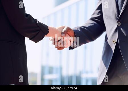 Stretta di mano di giovani partner commerciali interculturali in abiti in piedi fronte della telecamera contro l'architettura moderna mentre si salutano a vicenda Foto Stock