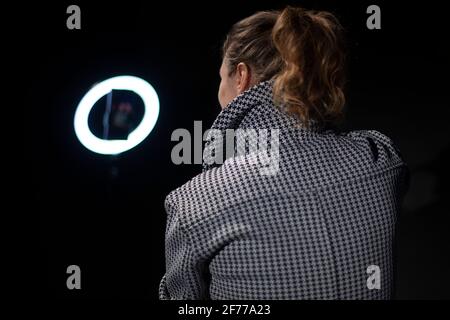 La ragazza rimuove il blog. La ragazza usa una lampada rotonda per illuminare il viso. Ripresa di un video blog con luce artificiale. Una donna in una giacca e wit Foto Stock