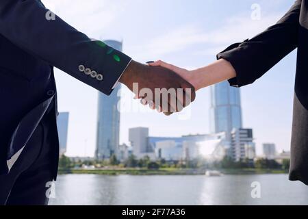 Stretta di mano di giovani partner commerciali interculturali salutano l'un l'altro mentre in piedi davanti alla macchina fotografica contro il paesaggio urbano e il lungofiume Foto Stock