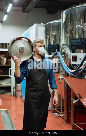 Giovane birrificio maschile operaio in maschera protettiva che porta cisterna sopra spalla mentre si muove lungo grandi serbatoi di acciaio in stabilimento di produzione Foto Stock