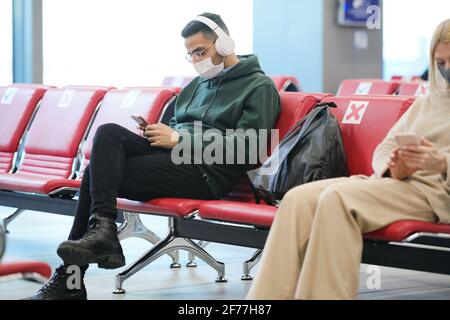 Giovane uomo con cuffie che ascolta la musica e scorre smartphone mentre si siede nella lobby dell'aeroporto moderno e si aspetta per il volo Foto Stock
