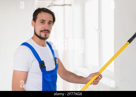 Colpo medio, riparatore in abiti da lavoro blu dipinge parete in appartamento Foto Stock