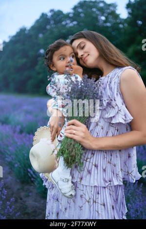 Vista laterale di una giovane madre sorridente che indossa un abito che porta un mazzo aromatico di bellissimi fiori viola. Donna che posa con la figlia piccola sulle mani nel campo di lavanda. Concetto di bellezza della natura. Foto Stock