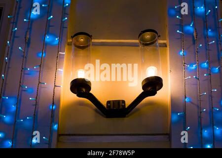 lampada da parete per illuminazione stradale in ferro con sfumature cilindriche in vetro e una lampadina elettrica sulla facciata di un edificio con un ghirland in blu, architettu Foto Stock