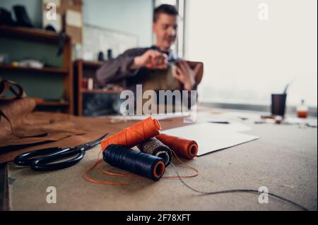 Diversi rocchetti di filo colorato sulla scrivania di un sarto, che è sullo sfondo in fuori fuoco cucire un prodotto in pelle. Foto Stock