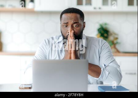 Triste frustrato afroamericano bearded uomo d'affari, freelancer o broker che lavora a distanza, seduto alla scrivania, indossando abiti eleganti, guarda ansiosamente al laptop, mani vicino al viso Foto Stock