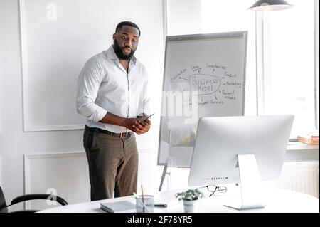 Allenatore d'affari afro-americano intelligente e autorevole, che conduce l'apprendimento a distanza o un briefing online per studenti o dipendenti, in piedi vicino a una lavagna a fogli mobili, guardando lo schermo di un computer Foto Stock