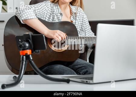 Giovane donna suona la chitarra acustica a casa per il pubblico online su laptop e smartphone. Corsi di formazione online per chitarra musica e formazione durante Foto Stock