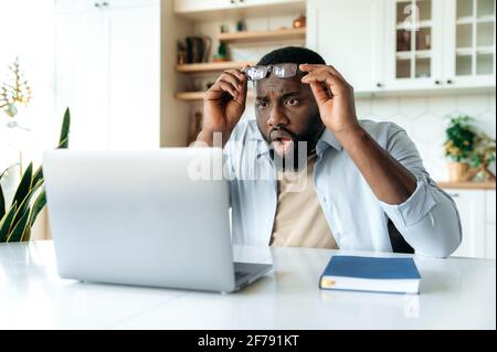 Scioccato uomo d'affari afro-americano, manager o broker, siede alla sua scrivania, guarda a sorpresa il suo laptop, legge notizie inaspettate, take off occhiali, espressione confuso del volto Foto Stock