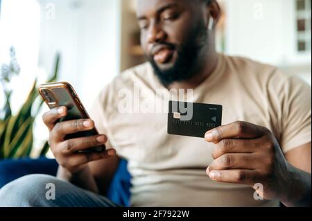 Difocuso giovane afro americano ragazzo bearded, in abiti casual, seduto sul divano a casa, tenendo un telefono cellulare e una carta di credito, pagare per gli acquisti online, inserendo un numero di carta Foto Stock