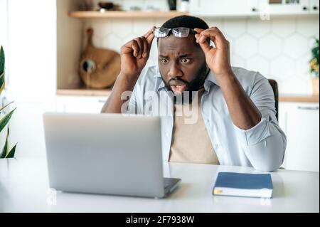 Sbalordito uomo bearded afro-americano, dirigente d'affari, freelance si siede alla sua scrivania, guarda il suo laptop a sorpresa, mentre togli gli occhiali, legge notizie o messaggi inaspettati Foto Stock