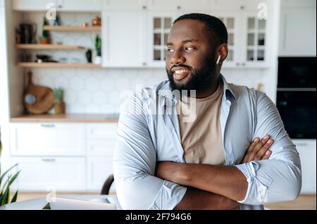 Ritratto di un giovane gioioso e sicuro afroamericano con barba che indossa cuffie in piedi in salotto, indossando abiti eleganti, guardando lateralmente, braccia incrociate davanti a lui, sorridente Foto Stock