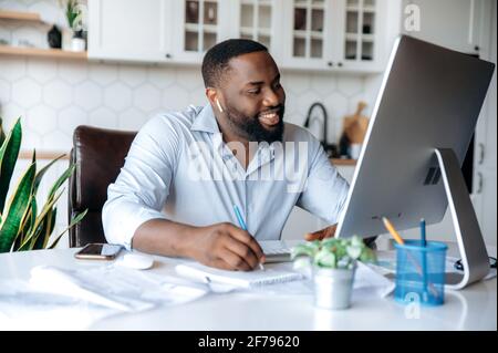 Sicuro riuscito bello giovane uomo d'affari afroamericano, freelancer o agente di bene immobile che lavora a distanza al calcolatore, che parla con il dipendente o il cliente via la chiamata di conferenza, sorridente amichevole Foto Stock