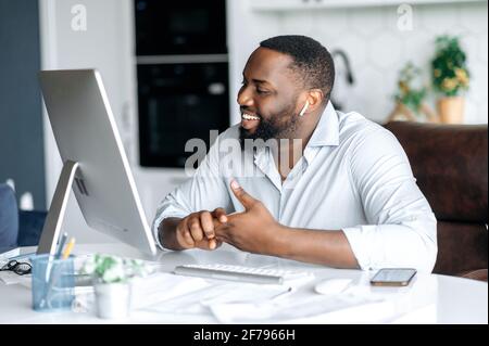 Gioioso e piacevole uomo afro-americano di successo, avvocato o agente immobiliare che lavora in remoto al computer, parlando con un collega o un cliente tramite videochiamata utilizza cuffie wireless, sorridente amichevole Foto Stock