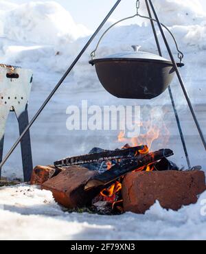 Sopra il fuoco appende una pentola in cui cucinare il cibo. Su un gancio su un treppiede, il vapore esce dalla padella. Inverno Camping cucina all'aperto Foto Stock