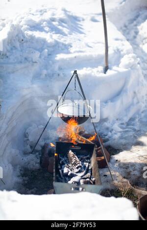 Sopra il fuoco appende una pentola in cui cucinare il cibo. Su un gancio su un treppiede, il vapore esce dalla padella. Inverno Camping cucina all'aperto Foto Stock