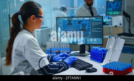 Donna tecnologa che fa un test di laboratorio che esamina un pallone con campione di sangue, chimico che tiene la provetta con i liquidi all'interno. Scienziato che lavora con vari tessuti batterici e immagini di scansione del DNA su computer Foto Stock