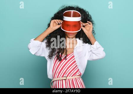 Giovane ragazza nera alla moda con un picco rosso trasparente o. hat che si sbirciano attraverso la visiera della telecamera in una cima a righe rosse e bianche su un turchese Foto Stock