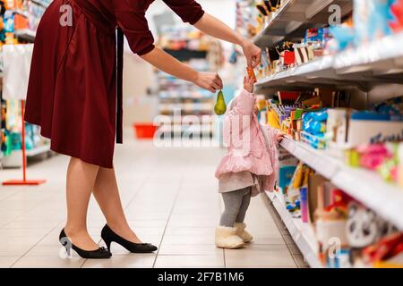 Una giovane madre dà il suo bambino, in piedi allo scaffale nel supermercato, una pera e una caramella. Primo piano. Il concetto di shopping familiare. Foto Stock