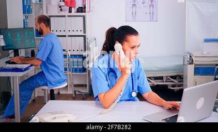 Assistente chirurgo che discute con il paziente al telefono dall'ospedale circa la diagnosi, infermiere maschio che lavora in background. Medico sanitario, medico receptionist che aiuta con la comunicazione di teleshealthcare Foto Stock
