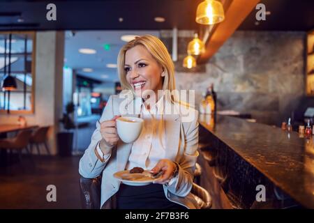 La donna bionda, felice e accattivante, si siede in un caffè del famoso hotel e si gode il suo caffè pomeridiano. Pausa, bevanda calda Foto Stock