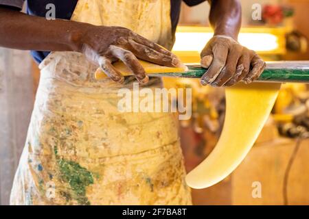 Uno Shaper da surf di un artigiano africano che lavora in un'officina di riparazione Con carta vetrata Foto Stock