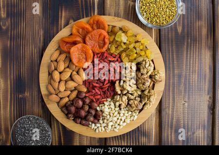 Albicocche secche, uvetta, bacche di goji, noci diverse, semi di chia e polline di api sul bordo rotondo da taglio sullo sfondo di legno marrone.Vista dall'alto. Foto Stock