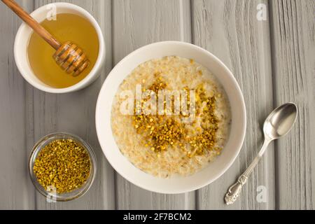 Colazione: Farinata d'avena con polline d'api e miele nella ciotola bianca sullo sfondo grigio di legno. Vista dall'alto. Foto Stock