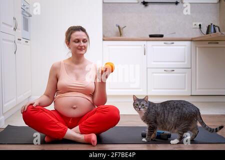 Una donna incinta si esercita a casa e tiene un frutto in mano come simbolo di cibo sano durante gravidanza Foto Stock