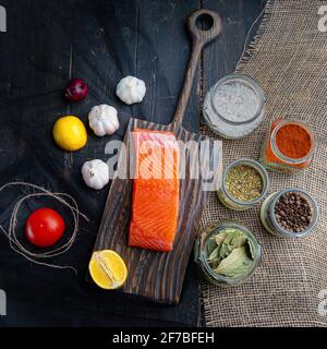 Un pezzo di salmone affumicato a freddo su un tavolo da servizio. Verdure e condimenti per il pesce. Sfondo di legno scuro. Foto Stock
