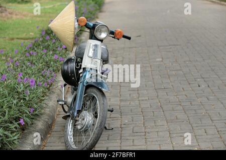 Ho Chi Minh City, 19 marzo 2021: Vecchio, usato, classico cuso 50 moto retrò impiccato con cappello a cono asiatico e casco parco su cemento pavement blocco. Foto Stock