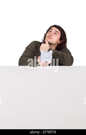 Pensieroso giovane uomo, che tiene una banca di soldi piggy, in piedi dietro un banner vuoto con spazio di copia per i messaggi e la pubblicità. Concetto finanziario, rais Foto Stock
