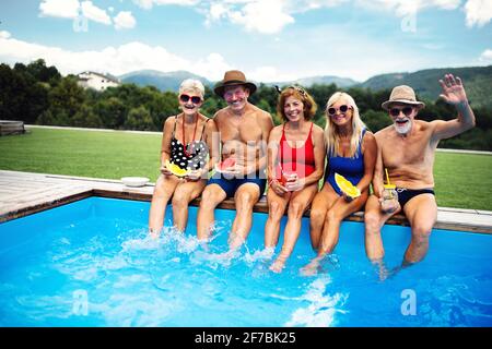 Gruppo di anziani allegri seduti accanto alla piscina all'aperto nel cortile, guardando la macchina fotografica. Foto Stock