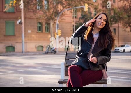 gioiosa giovane donna seduta su una panchina di strada ridendo mentre fa una chiamata telefonica, concetto di comunicazione e stile di vita urbano, copyspace per il testo Foto Stock